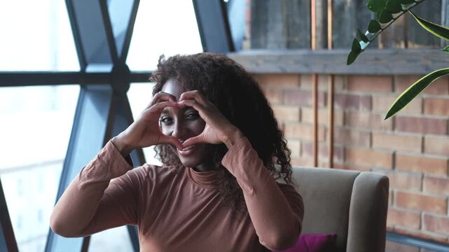 Portrait of a cute African American woman with a beautiful smile, with long curly hair. A woman looks at the camera and shows a heart with her fingers, a symbol of love.
