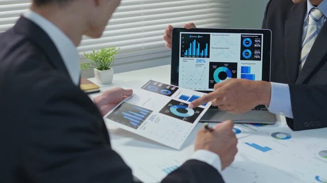 Business professionals discussing and explaining data charts on a laptop screen during an office meeting, with reports and documents on the desk, representing teamwork, analysis and strategic planning