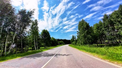 Naklejka premium Curved asphalt road surrounded by lush green trees under a blue sky with scattered clouds