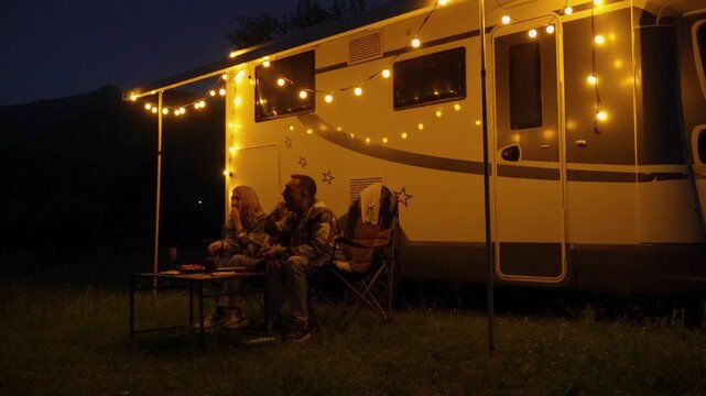 Romantic couple relaxing and talking outside their campervan under the warm glow of string lights during a beautiful summer evening. Perfect vacation getaway for two in nature