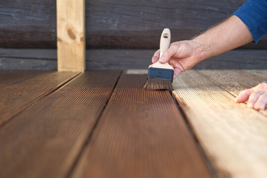 Brown oil for protection of wood by staining. Painting a larch for protecting and pests. Woodworker applying finish to terrace deck board. Repairman varnishing with brush wooden plank floor surface.