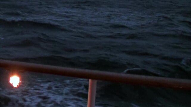 Witness Arctic Ocean's profound darkness from ship's deck. Turbulent, cold waves surge past sturdy rail, where solitary, gentle light casts warm glow upon frothing sea during twilight passage.