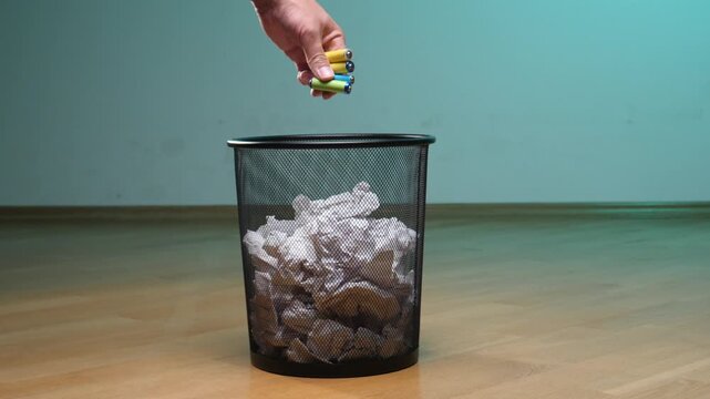 Hand dropping used aa batteries into a mesh trash can filled with crumpled paper, highlighting the improper disposal of hazardous waste and the importance of recycling for sustainability