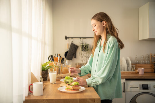 Young caucasian woman in green shirt preparing a healthy fresh salad and sandwich in the kitchen. Beautiful female cooking morning breakfast with fresh vegetables at wooden kitchen counter.