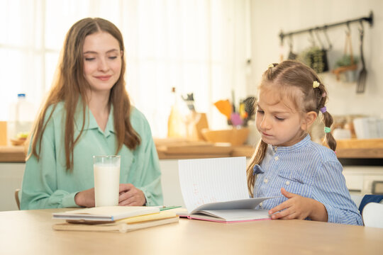 Little schoolgirl flipping pages of her notebook with a glass of milk on the table. Focused daughter reading book or notebook with mother watching her at kitchen desk.