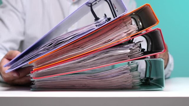 Person carrying a tall stack of colorful, full ring binders, representing a heavy workload, bureaucracy, and extensive paperwork in an office environment