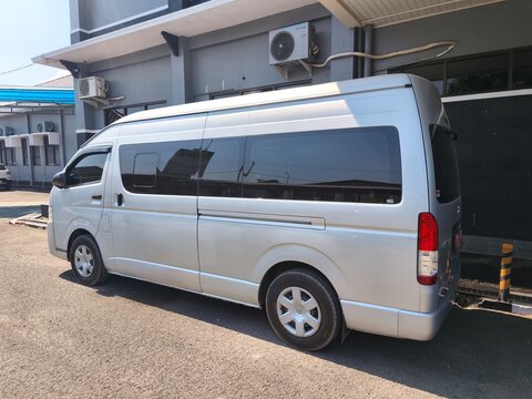 A silver Toyota HiAce commuter passenger van parked outside an office building under the canopy roof during a sunny day.