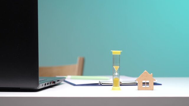 Laptop, clipboard, hourglass, and a small wooden house model on a white desk, illustrating real estate investment, home loan application deadlines, and property acquisition time limits