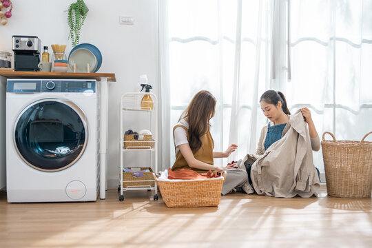 Woman laundry basket washing machine clothing home interior housework fabric window, two women sorting clothing on floor in bright laundry room with wicker basket, calm domestic chore scene
