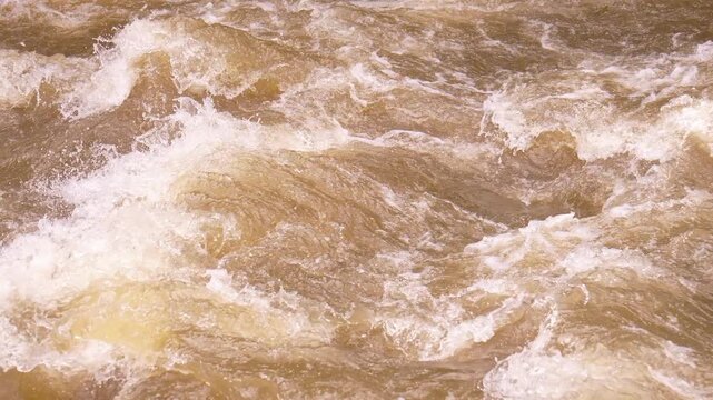 Detail shot of turbulent muddy water with white foam from a powerful river current after heavy rainfall, showing the impressive and dangerous force of nature during a flood event slow motion