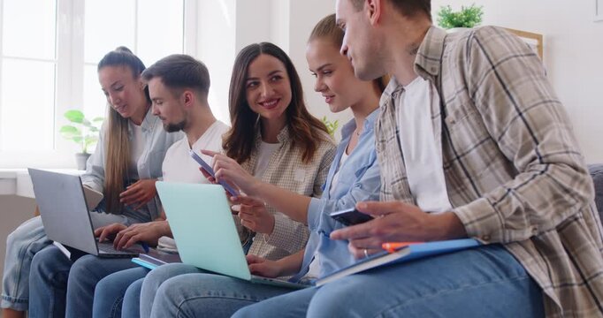 Portrait of a group of young people friends or students in casual clothes sitting in a row on sofa at home and using laptops and mobile phones together watching news in social media. 4k video.