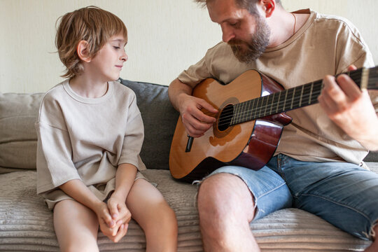 Loving father with beard playing acoustic guitar for son sitting on couch. Father-son bonding, family values, authentic home life atmosphere. Selective focus.