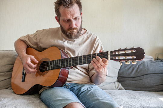Focused bearded man sitting on sofa and playing classical acoustic guitar at home. Man wearing casual beige t-shirt and jeans, practicing music. Creative leisure and personal fulfillment.