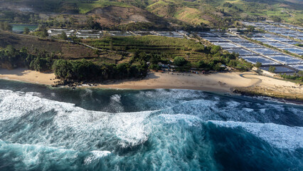 Drone aerial view of a tropical beach bay with breaking ocean waves, sandy shoreline, and rolling green hills with farmland in the background under warm daylight. © Fredy