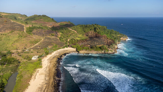 Wide aerial view of a tropical coastline in Southeast Asia, showing forested cliffs, terraced hillside farmland, a small sandy cove, and turquoise waves meeting the deep blue ocean.
