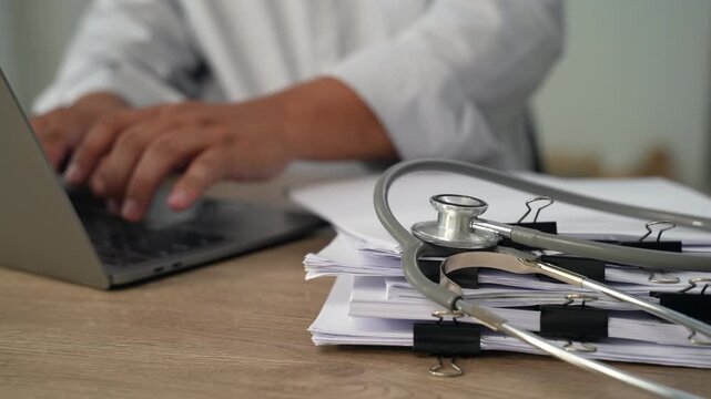 medical paperwork beside a laptop and stethoscope on a wooden desk, documenting information on stacked forms for an organized healthcare records workflow.