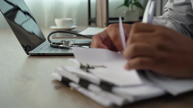 medical paperwork beside a laptop and stethoscope on a wooden desk, documenting information on stacked forms for an organized healthcare records workflow.
