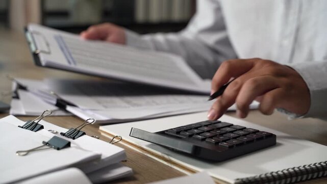 Project management  person using a calculator while reviewing printed documents on a desk, showing calculation and document checking for accounting, expense review, and financial reporting.