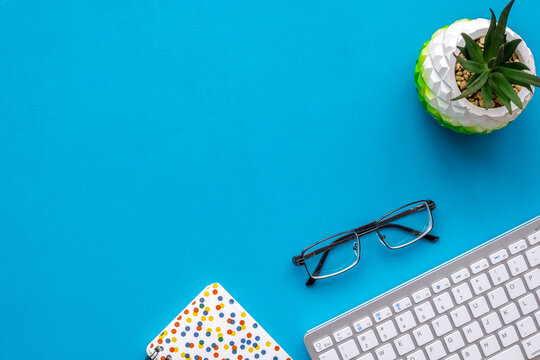 Work desk of manager. Computer keyboard, glasses, stationery, plant on blue background top view copy space