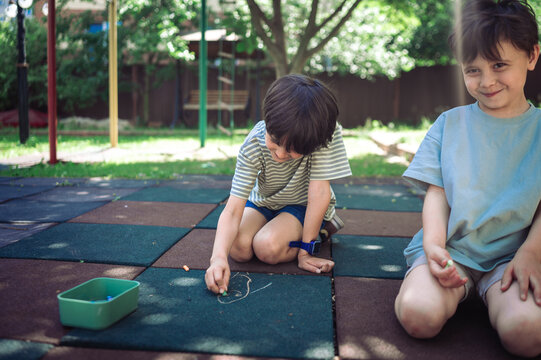 A boy draws with chalk while his brother sits on the playground