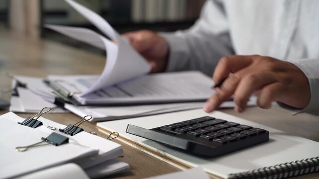 Project management  person using a calculator while reviewing printed documents on a desk, showing calculation and document checking for accounting, expense review, and financial reporting.
