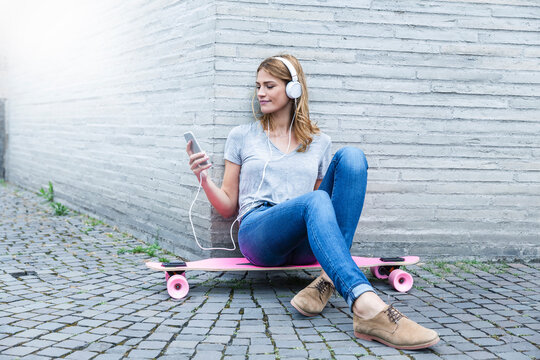 Germany, Cologne, young woman sitting on pink skateboard hearing music with headphones