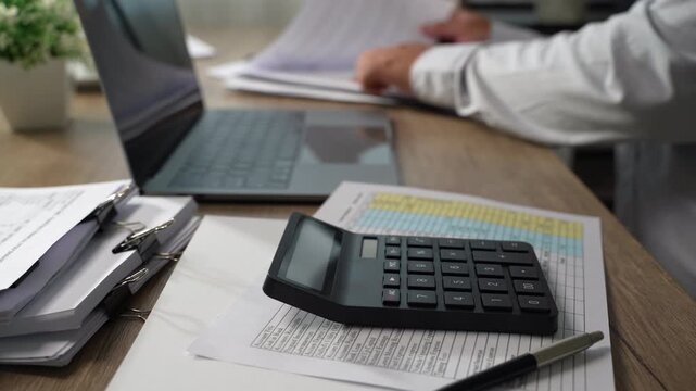 Project management  person using a calculator while reviewing printed documents on a desk, showing calculation and document checking for accounting, expense review, and financial reporting.