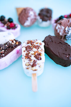 Colorful assortment of homemade yogurt popsicles. Vanilla and chocolate decorated with nuts and frozen berries on ice blue background. Perfect for summer party. Vertical top view close-up with short d
