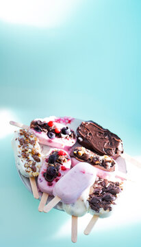 Colorful assortment of homemade yogurt popsicles. Vanilla and chocolate decorated with nuts and frozen berries on ice blue background. Perfect for summer children's birthday party. Vertical overhead c