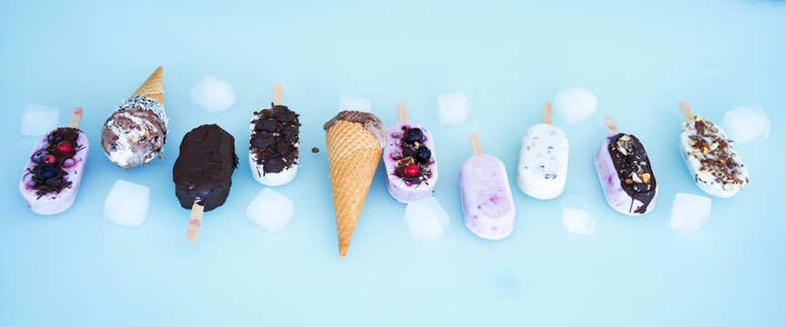 Colorful row of homemade yogurt and milk ice pops. Vanilla and chocolate decorated with nuts and frozen berries with ice cubes on bright blue background. Perfect for a summer children's birthday party