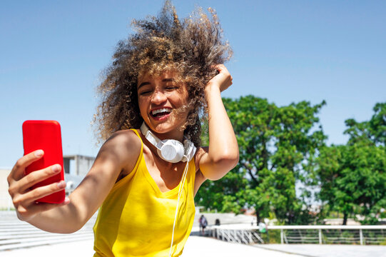 Young woman with hand in hair laughing while taking selfie through smart phone