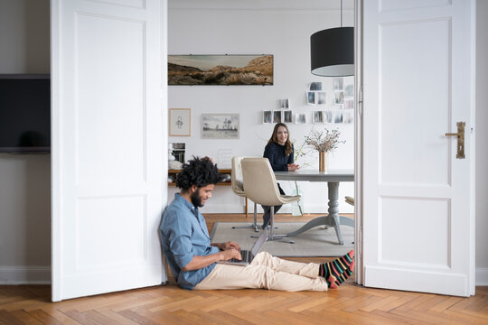 Man at home sitting on floor working with laptop with woman in background