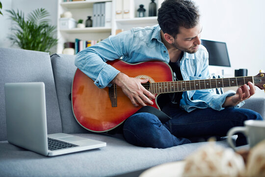 Young man at home sitting on couch playing guitar next to laptop