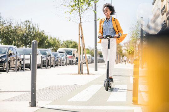 Happy young woman with earphones riding e-scooter in the city