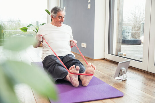 Senior man pulling resistance band watching online tutorial through tablet PC at home