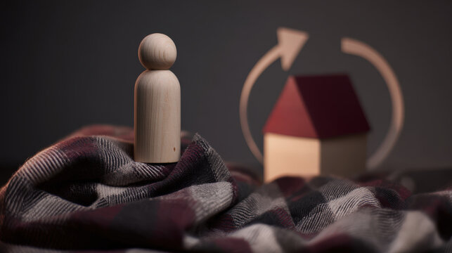 A solitary wooden peg doll sits on a checkered blanket, with a miniature house and an arrowed circular symbol in the background, suggesting themes of home and cycles.