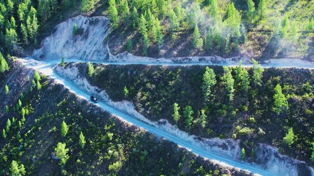 Dark SUV descending lower switchback on pale gravel mountain road through steep evergreen hillside under bright oblique sunlight and rugged forest landscape, with road, gravel, mountain, forest detail