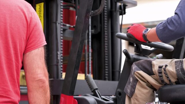 Forklift operator driving heavy machinery while worker guides loading process in warehouse. Industrial safety warning stickers visible on lifting mast. High quality 4k footage