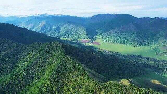 Bird's-eye panorama of forested mountain ridgelines descending into sunlit valley with patchwork farm fields, scattered rural buildings, distant peaks, and expansive horizon