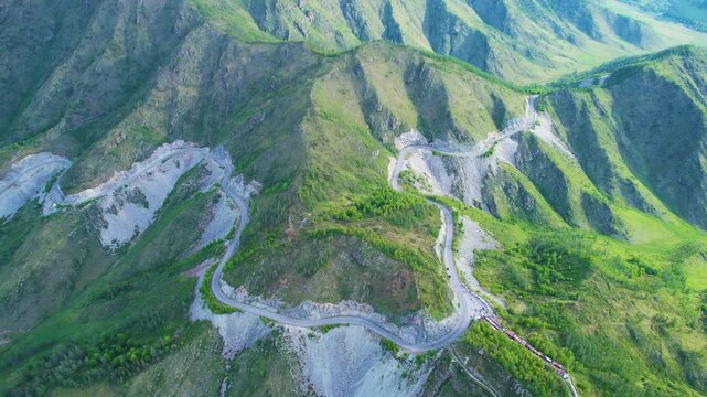 High aerial view of winding mountain road with hairpin switchbacks crossing grassy ridges, rocky slopes, valley contours, remote alpine landscape and visible vehicles