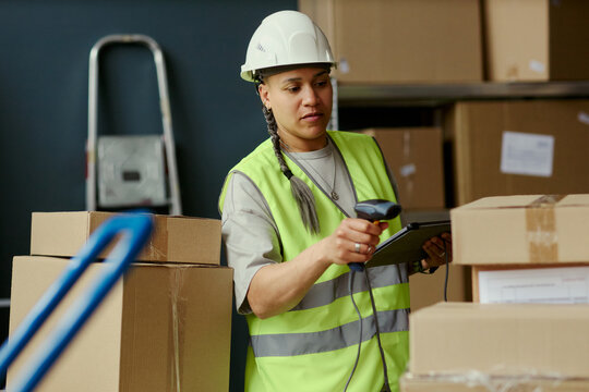 Medium shot of young adult biracial woman scanning packages with barcode reader and checking tablet in warehouse, managing inventory and fulfillment. Ideal for logistics, supply chain, ecommerce