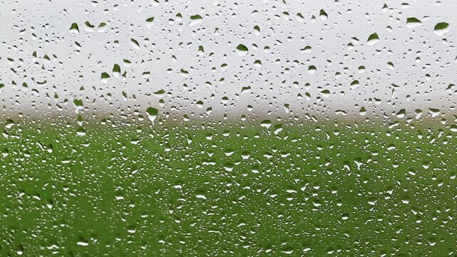 Raindrops on glass surface with blurred green field and overcast sky in background, natural water texture, wet weather mood