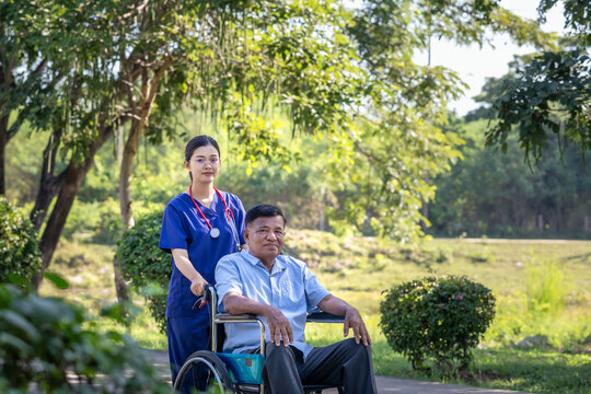 Elderly man patient in a wheelchair being cared for by a medical professional. Asian nurse take care senior male sit on wheelchair in park, outdoor.