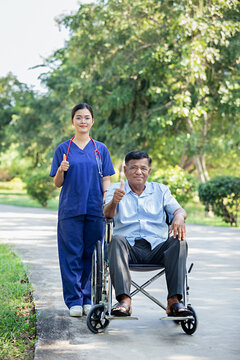 Female nurse cares for elderly man sitting in wheelchair in park. Positive image depicting dedicated professional care for an elderly patient in a natural outdoor setting.