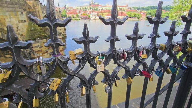 Close-up of love locks hanging on an ornate iron fence near Charles Bridge in Prague, with the Vltava River, old stone bridge, red rooftops, and Prague Castle blurred in the background.