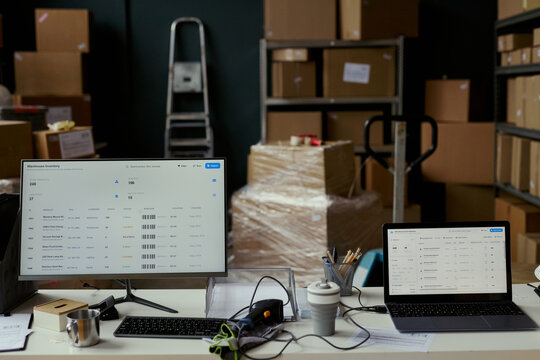 Wide shot of warehouse workstation displaying inventory management dashboard on desktop monitor and laptop beside packed boxes. Supporting ecommerce logistics, stock control, fulfillment marketing