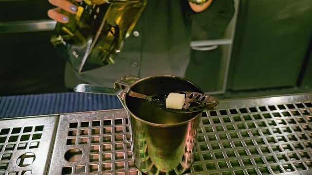 Close-up of traditional absinthe preparation at a cocktail bar, with sugar cube on a slotted spoon, metal cup, green bottle, stainless drip tray, and moody nightlife lighting.