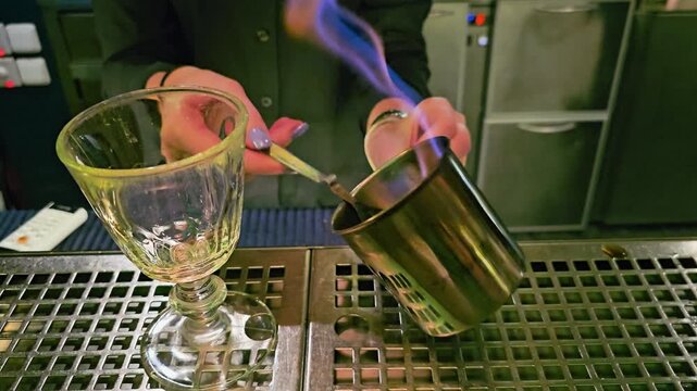 Bartender performs classic absinthe ritual with flaming sugar cube on a perforated spoon, metal mixing cup, empty glass goblet, green bar lighting, and moody lounge atmosphere indoors.