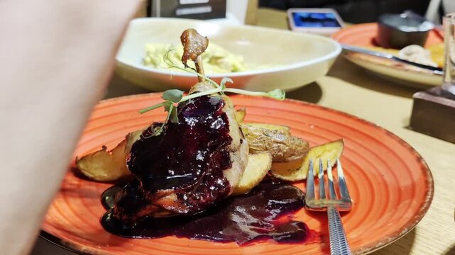 Duck confit dish with rich dark berry sauce, crispy potato wedges, and delicate microgreens on an orange plate in restaurant setting, ready for consumption