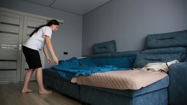 Young woman making bed in contemporary living room, spreading blue sheet over beige mattress pad on blue sofa, preparing for sleep or tidying up home interior.